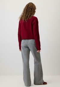 Kobieta z kręconymi włosami stoi tyłem, ubrana w czerwoną swetrową bluzę i szerokie szare dżinsy, na tle gładkiego białego tła.