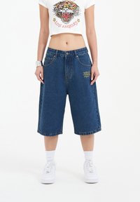 Dark blue denim shorts, knee-length, with a relaxed fit. Features yellow embroidered text and classic five-pocket design. White sneakers complete the look.