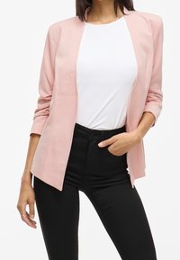 Růžový blazer s hladkou strukturou a otevřeným předním designem, v kombinaci s bílým topem a černými džínami s vysokým pasem. Střih na míru a strukturované rukávy.