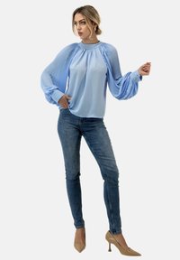 Lichtblauwe blouse met geplooide mouwen, ronde halslijn en manchet; gecombineerd met een strakke blauwe jeans en beigekleurige hoge hakken.