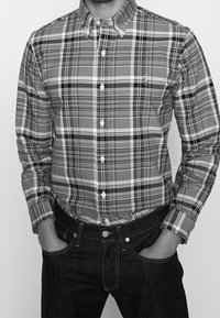 Red and black plaid button-up shirt with a collar, white buttons, and long sleeves. Worn with dark jeans, hands in pockets.