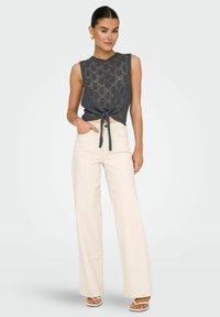 Haut sans manches en dentelle grise avec un détail de nœud à l'avant, associé à un pantalon beige taille haute et large ainsi qu'à des sandales à bout ouvert.