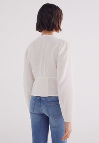 Weiße strukturierte Bluse mit langen, gepufften Ärmeln und einer taillierten Taille, kombiniert mit blauen, figurbetonten Jeans. Die Rückansicht zeigt ein schlichtes Design und klare Linien.