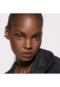 Gros plan sur un portrait d'une personne à la peau foncée, présentant une peau lisse, une lèvre brillante, des sourcils définis, et une boucle d'oreille argentée surdimensionnée. Drapé de tissu noir.