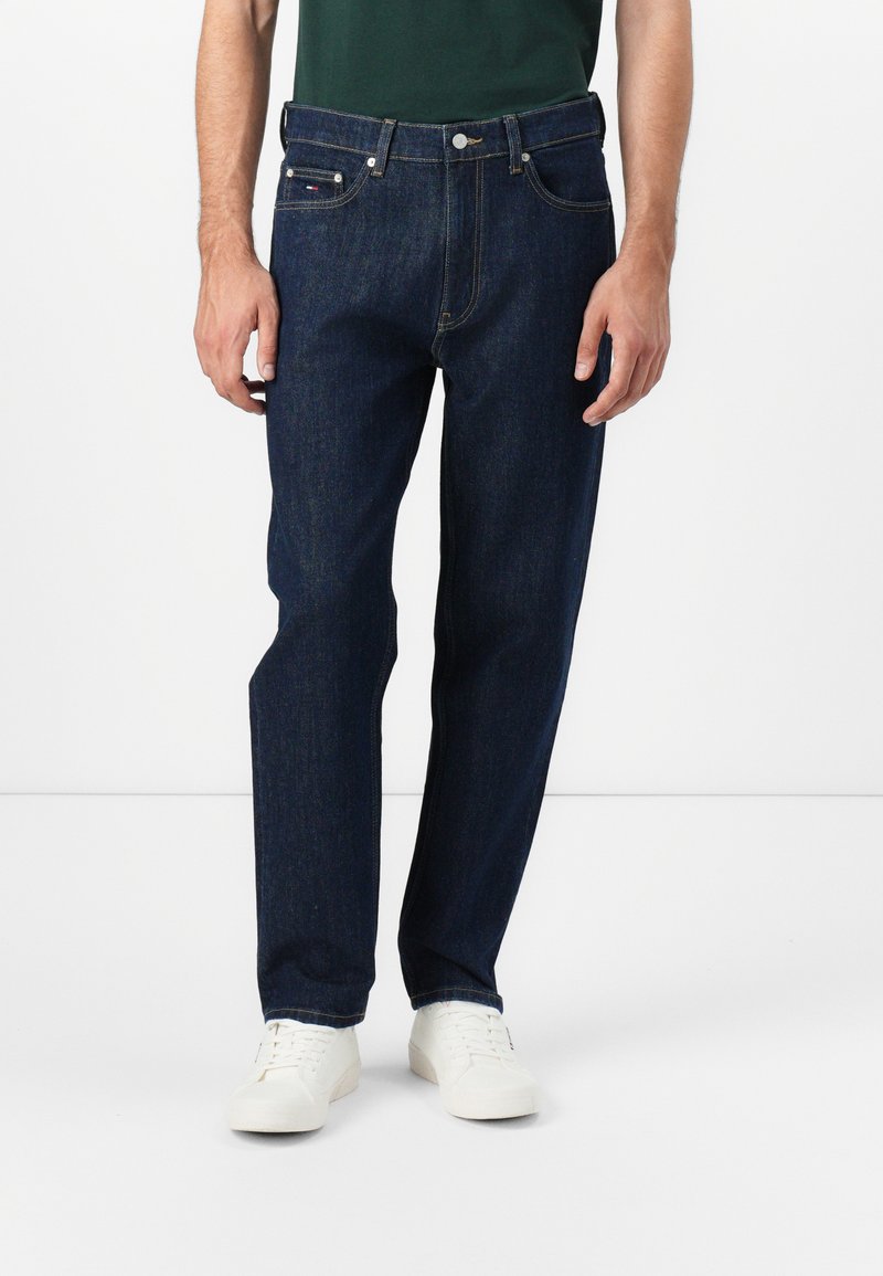 Dunkelblaue Jeans aus Denim mit geradem Bein, fünf Taschen und silbernem Zubehör. Getragen mit einem grünen Hemd und weißen Sneakers.
