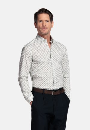 Mann trägt ein weißes gemustertes Hemd mit Knöpfen, schwarze Hose und braunen Gürtel, steht mit den Händen in den Taschen vor einfarbigem Hintergrund.