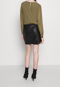 Top verde oliva con maniche texturizzate, abbinato a una mini gonna di pelle nera. Stivali neri alla caviglia con tacco alto completano l'outfit.