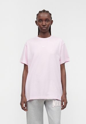 Vrouw met ingevlochten haar die een lichtroze Nike T-shirt en lichtgrijze Nike joggingbroek draagt, staand tegen een effen achtergrond.