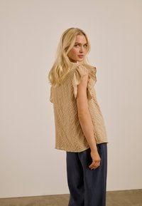 Femme blonde jetant un coup d'œil par-dessus son épaule, portant un haut sans manches beige texturé avec des manches à volants et un pantalon ample bleu foncé sur un fond uni.