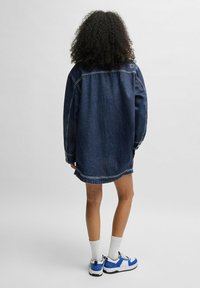 Donkerblauwe denim jurk met een ontspannen pasvorm, voorzien van een gebogen zoom en gestikte accenten. Gecombineerd met wit-blauwe sportschoenen.
