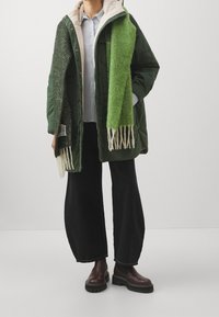 Grön puffjacka med en beige pälskantad huva, tillsammans med en grön halsduk med vita fransar. Modellen bär vida svarta byxor och bruna stövlar.