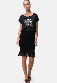 T-shirt noir avec imprimé tropical doré ; assorti à une jupe en crochet noir avec un ourlet à franges. Le modèle porte des sandales noires.