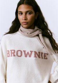 Suéter crema con "BROWNIE" bordado en rosa, combinado con una bufanda marrón claro. Textura suave, ajuste relajado, cuello y puños acanalados.