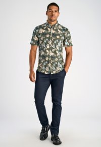 Camicetta a maniche corte con bottoni in verde, con fiori crema e blu. Indossata con pantaloni blu navy e scarpe nere.