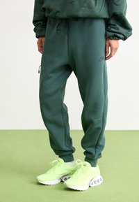 Osoba stojící na světle zeleném koberci u bílé stěny, oblečená v tmavě zelených teplácích Nike a jasně neonově zelených teniskách.