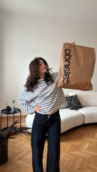 Braune Papiertüte mit schwarzem "Sandro"-Logo, getragen mit einem weißen und marineblauen, gestreiften Langarmshirt und dunklen Jeans aus Denim.