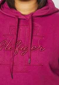Fioletowa bluza z kapturem z haftowanym logo i sznurkiem. Miękki materiał o gładkiej teksturze. Zawiera akcenty z metalowymi elementami w kolorze srebrnym.