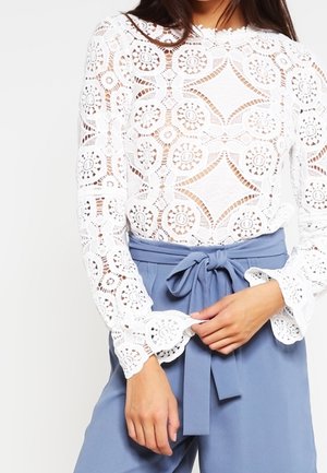 Top blanc en dentelle à manches longues avec motifs floraux, associé à un pantalon bleu taille haute noué d'un nœud à la taille.