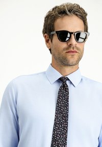 Camicia azzurra chiara con colletto a punta, abbinata a una cravatta scura con un motivo a paisley e occhiali da sole scuri.