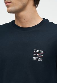 Νεαρός που φοράει σκούρο μπλε T-shirt με μικρό λογότυπο Tommy Hilfiger και το κείμενο "Tommy Since 1985 Hilfiger" στο στήθος.