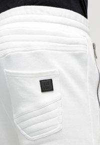 Bílé joggingové kalhoty s texturovanou kapsou a černou etiketou s nápisem "BLACK KAVIA." Mají hladký povrch a design žebrovaného pasu.