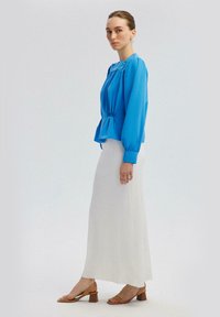 Blusa blu a maniche lunghe con dettaglio in vita elasticizzato, abbinata a una gonna bianca fluida e plissettata e sandali marroni con tacco a blocco.