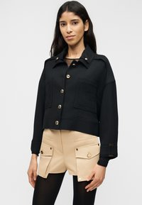 Γυναίκα που φοράει μαύρο cropped σακάκι με χρυσά κουμπιά πάνω από μπεζ σορτς με μεγάλες μπροστινές τσέπες και μαύρες καλτσοδέτες, στέκεται μπροστά σε απλό φόντο.