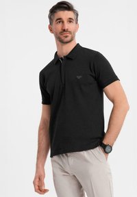 Černé polo tričko s krátkým límcem, přiléhavý střih, decentní logo na hrudi, v kombinaci s světlými kalhotami a tmavými hodinkami.