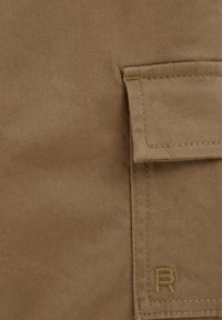 Beige stof met een twill-textuur, voorzien van een pocketflap en een geborduurd "R"-logo rechtsonder. Stiksel in een contrasterende kleur.