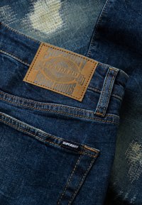 Donkerblauwe denim jeans met een gestikte leren patch met de tekst "Superdry Standard Denim Goods" en een klein zwart stoffen merkteken.