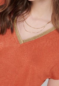 Kobieta ubrana w pomarańczowy sweter z złotymi wykończeniami przy dekolcie w kształcie litery V oraz dwoma delikatnymi złotymi naszyjnikami.