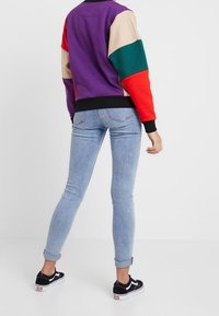 Sudadera multicolor con secciones moradas, rojas, verdes y beiges; puños y cinturilla negros. Usada con vaqueros ajustados de mezclilla azul claro y zapatillas negras.