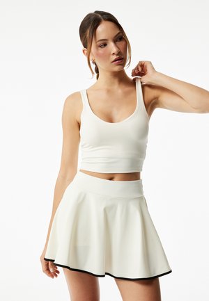 Jonge vrouw met bruin haar in een wit mouwloos crop topje en witte rok met zwarte bies, die het topbandje aanpast tegen een effen witte achtergrond.