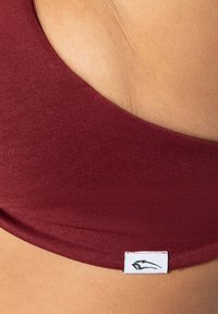 Bordeaux sportbh van gladde stof met een brede halslijn, voorzien van een klein wit label met een zwart logo aan de zijkant.