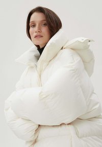 Duża biała pikowana kurtka puchowa z wysokim kołnierzem, pikowaną teksturą i zamkiem błyskawicznym. Tkanina wydaje się gładka i ocieplona.