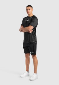 Camiseta deportiva negra con logo, combinada con pantalones cortos negros que tienen un acento de flecha blanca. El modelo lleva zapatillas blancas.