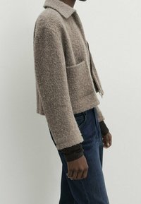Person som bär en texturerad taupe-färgad kort jacka med framfickor över en mörk långärmad topp och blå jeans, stående i profil.