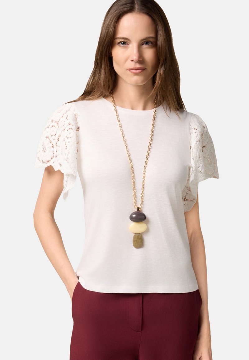 Haut blanc à manches courtes avec des manches accentuées de dentelle ; associé à un pantalon bordeaux. Un collier en chaîne doré avec trois pierres décoratives.