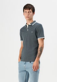 Grå polo skjorte med en kontrasterende lys krage, korte ermer og to-knaps stolpe. Har blå stripete detaljer ved ermeinnsnæringene.
