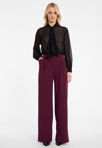 Vrouw met blond haar die een doorzichtige zwarte blouse met een strik en hoge taille wijde paarse broek draagt, staand met één hand in haar zak.