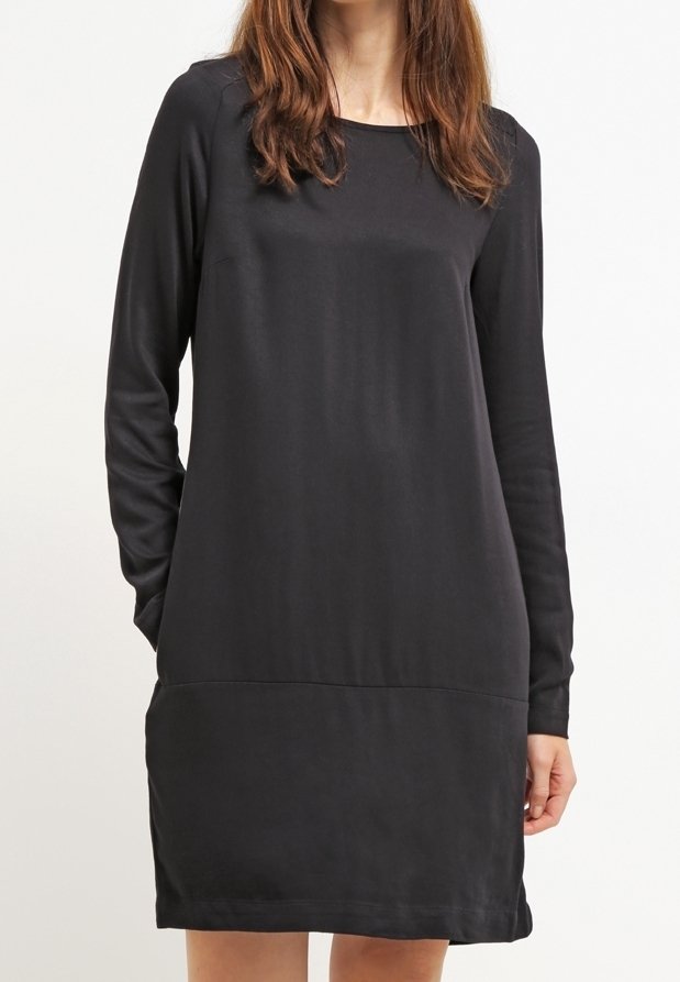 Femme portant une robe noire à manches longues, longueur genou, avec un col rond, debout devant un fond blanc uni.