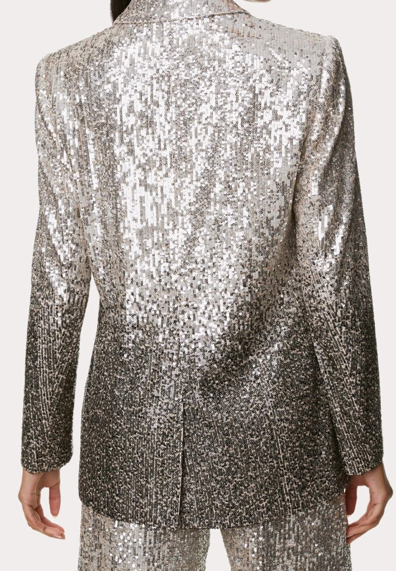 Vue arrière d'un blazer argenté à sequins avec manches longues et pantalon assorti porté par une personne.