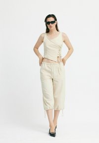 Beige 3/4-Hosen und ein ärmelloses Top mit Schnürdetails. Glatter Stoff mit einer leicht strukturierten Oberfläche. Schwarze spitze High Heels und Sonnenbrille.