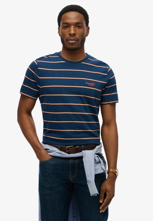 Marine gestreept t-shirt met oranje strepen, korte mouwen, ronde hals. "SuperDry" logo op de borst. Donkere jeans, bruine riem, licht shirt geknoopt om de taille.