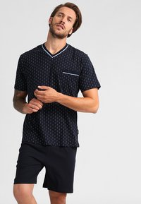 Marineblauw kortemouwen shirt met een V-hals, witte patronen en een borstzak. Gecombineerd met zwarte shorts van gladde stof en een comfortabele pasvorm.