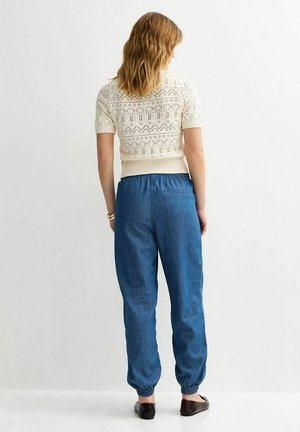 Femme aux cheveux blonds mi-longs portant un pull à manches courtes blanc à motifs, un pantalon de jogging bleu à taille élastique et des ballerines noires.