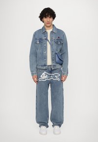 Giacca in denim e jeans con grafica cartoon bianca. Colore azzurro chiaro, vestibilità rilassata e un piccolo logo rosso sulla giacca. Sneakers bianche e borsa blu.