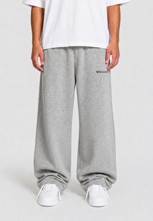 Person iført løstsiddende lysegrå joggingbukser med Colucci-logo, hvide sneakers og en hvid t-shirt med korte ærmer, stående på en ensfarvet baggrund.