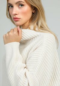 Pull en tricot crème, avec un col haut, des poignets côtelés et un motif texturé. Gros plan sur un modèle tenant légèrement le col.
