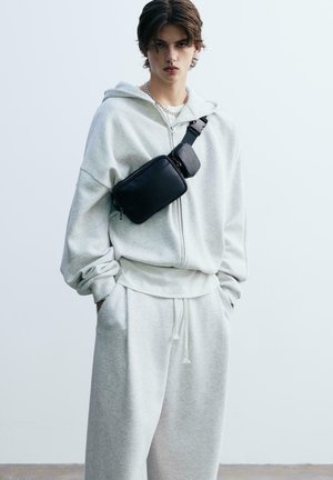 Grå, strukturerad huvtröja med dragkedja och framfickor, tillsammans med matchande sweatpants. En svart axelväska bärs över bröstet.